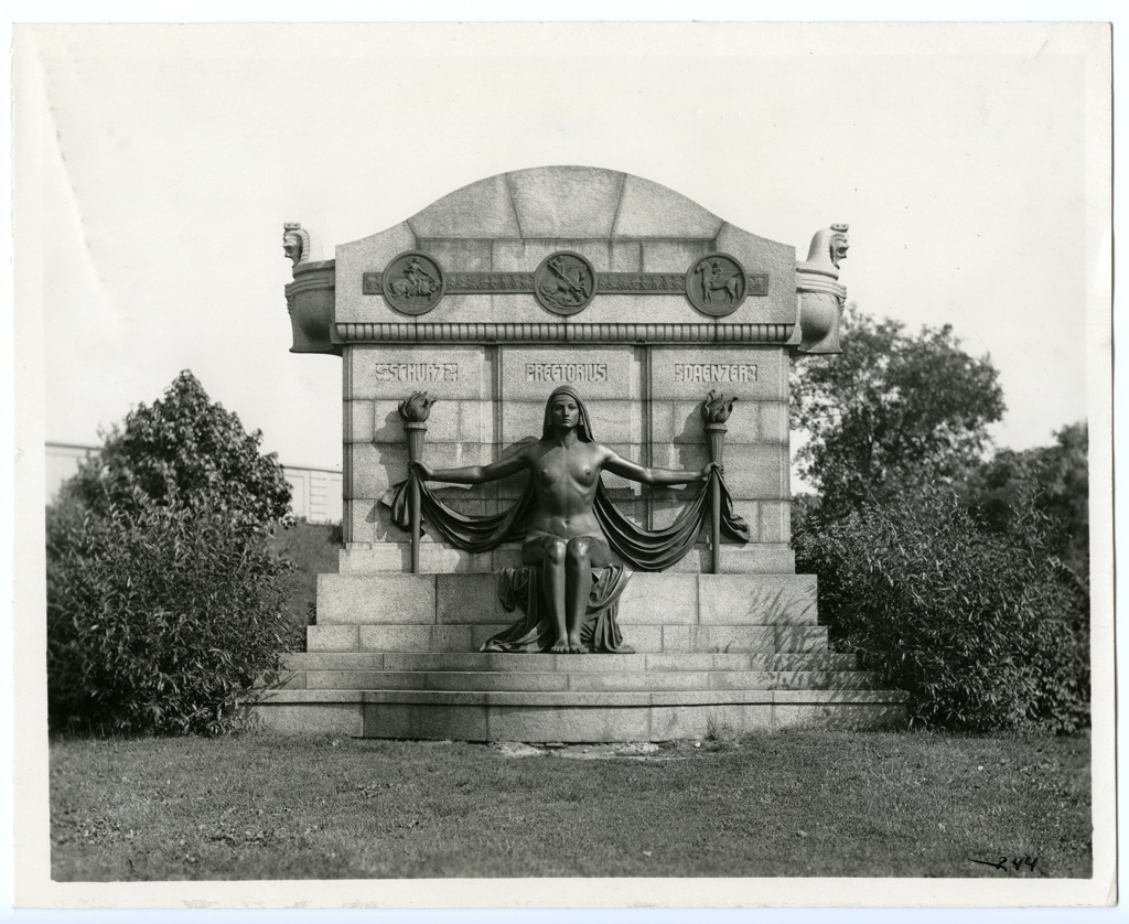"THE NAKED TRUTH." (PREETORIUS-SCHURZ-DAENZER MONUMENT). Horizontal, black and white photograph showing a monument with a statue of a naked woman seated holding a touch in each outstretched hand. Three names above her head read: "Schurz. Preetorius. Daenzer." On either side of the monument are shrubs.